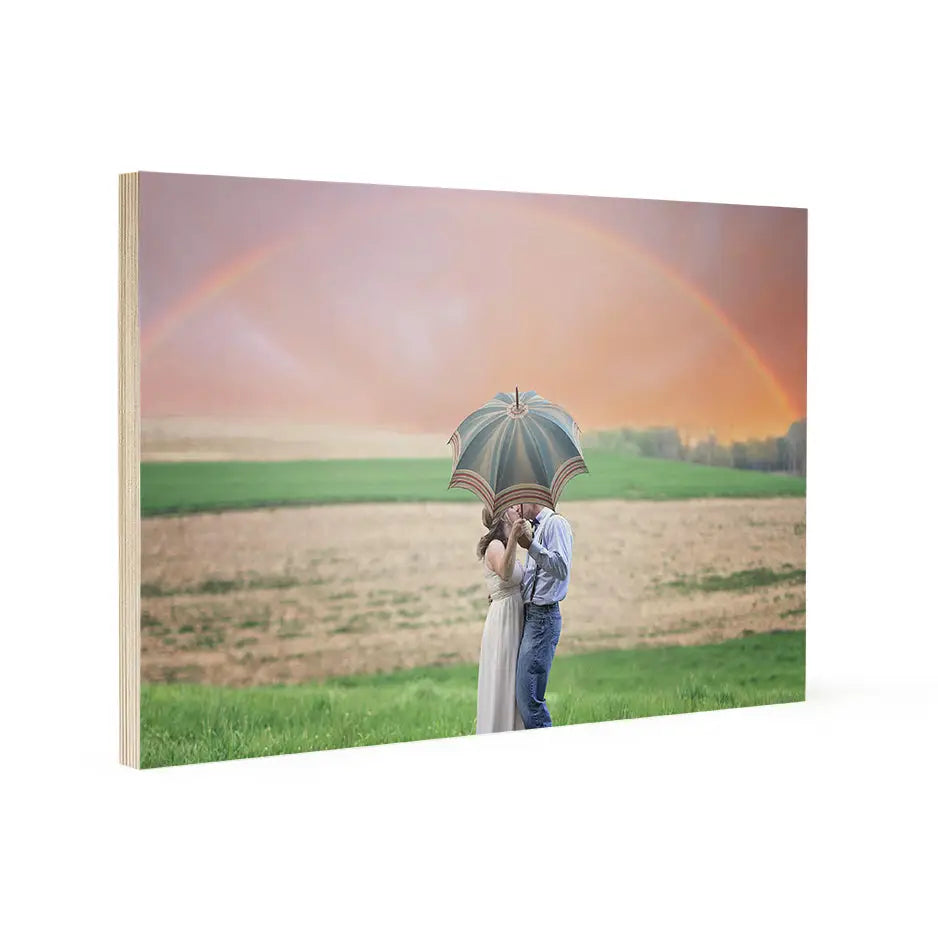 Couple embracing under an umbrella in a field with a rainbow near Woodsnap’s Birch Wood Prints.