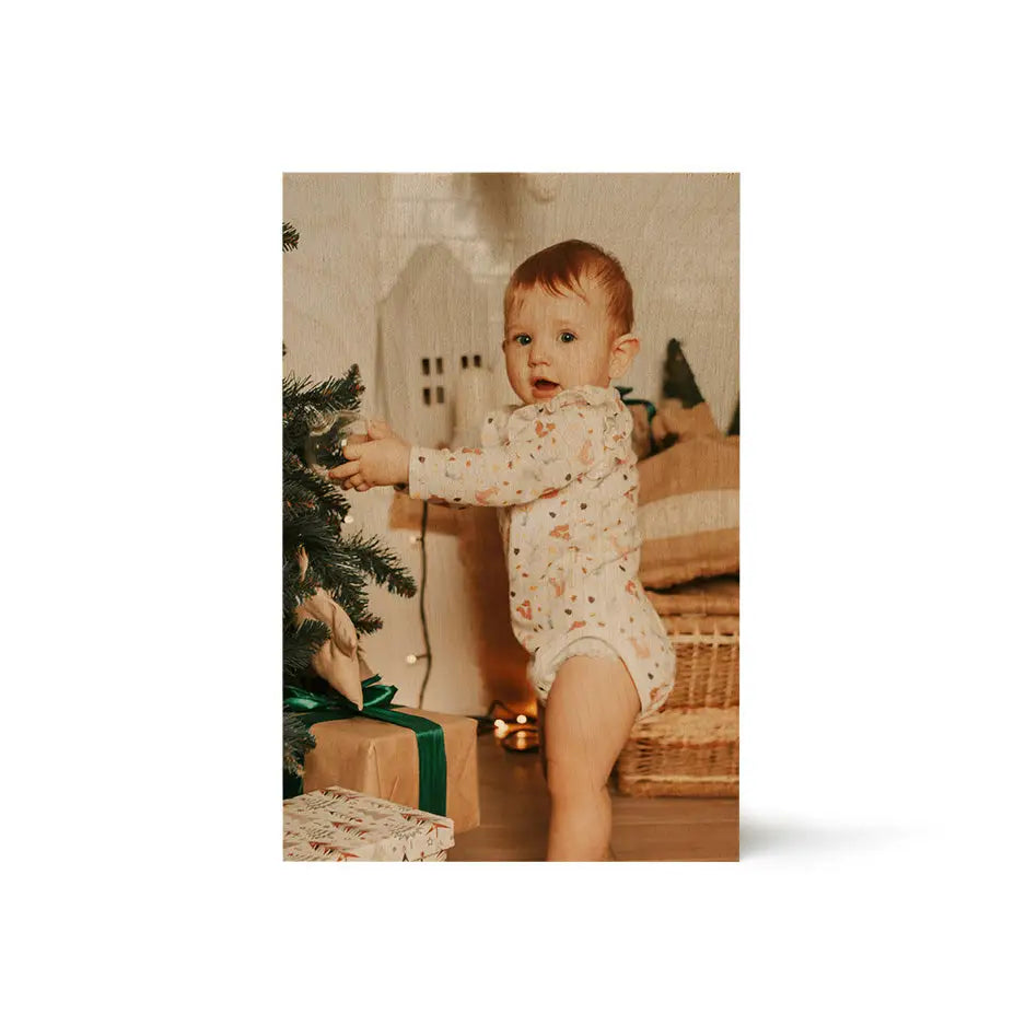 Baby in a white patterned onesie by a Christmas tree on a vintage matte wood print