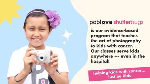 Smiling child with a digital camera and flower accessory supporting Pablove Foundation.