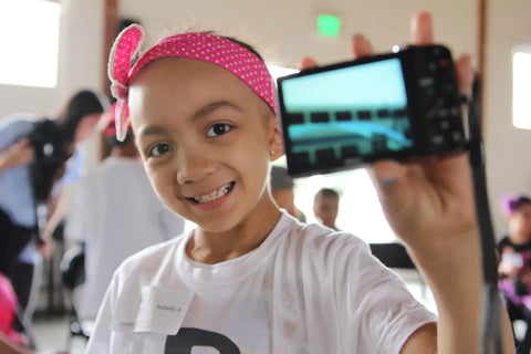 Smiling child with a pink headband holds a smartphone for Pablove Foundation event.